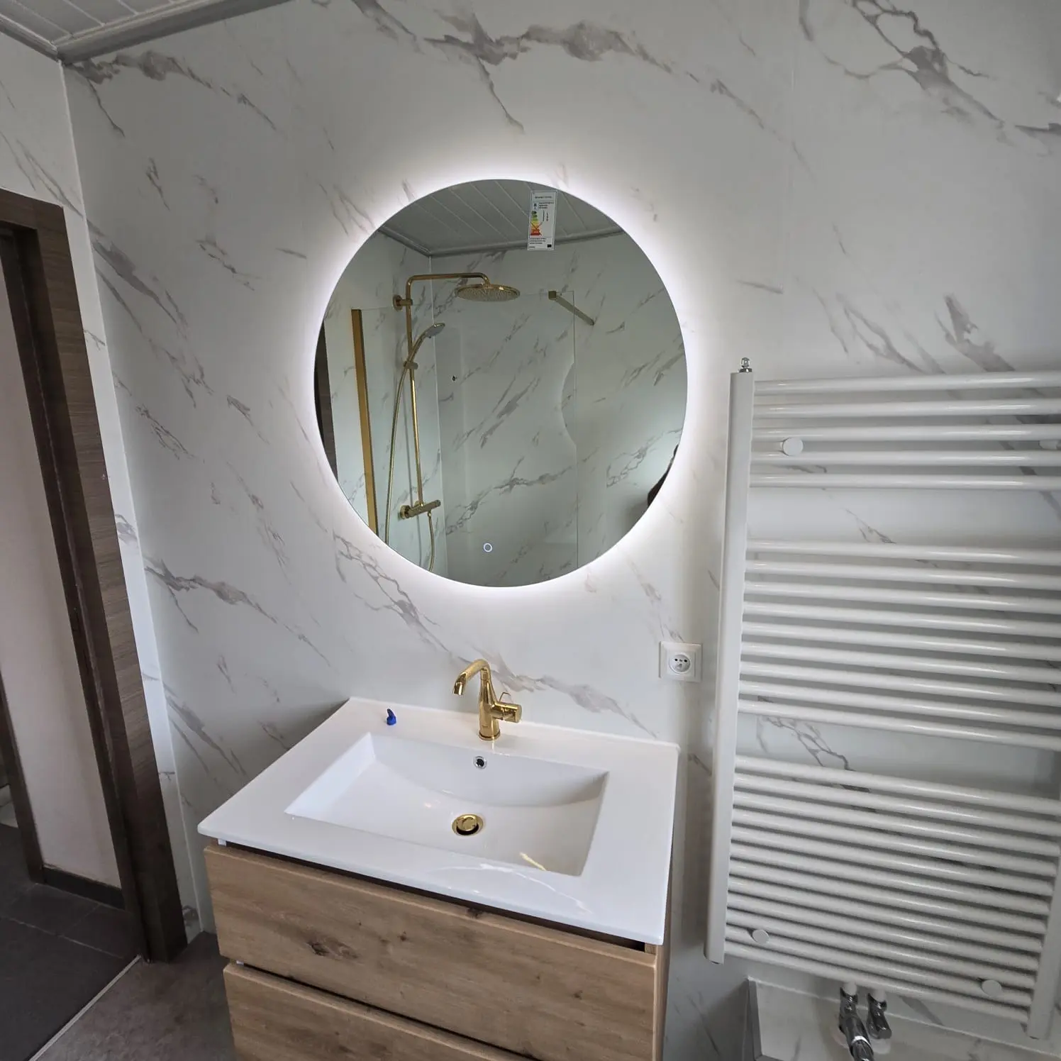 nieuwe badkamer met marmerlook wanden, ronde spiegel met verlichting, houten wastafelmeubel en gouden kraan. inloopdouche zichtbaar in spiegel.